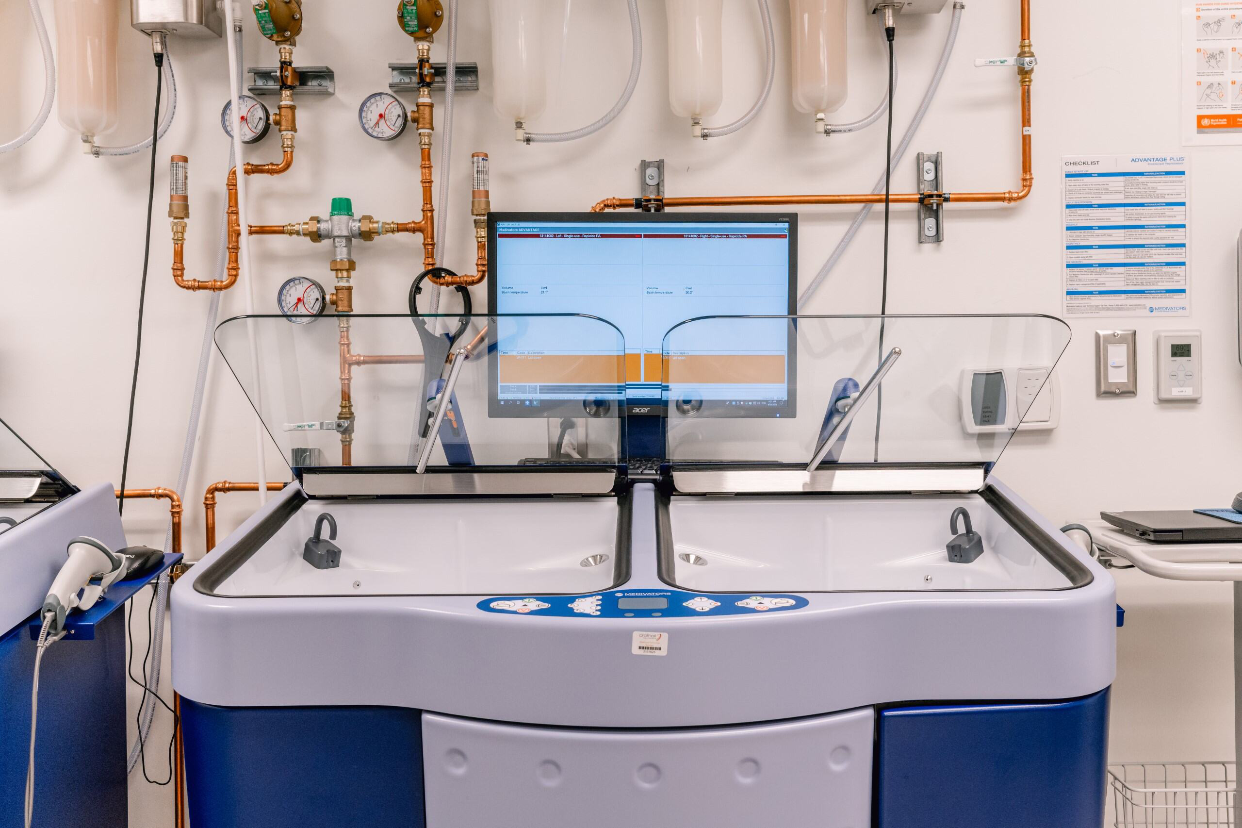 A modern medical device with twin basins, a central touchscreen showing data, pipes, gauges, and equipment in a clinical room.