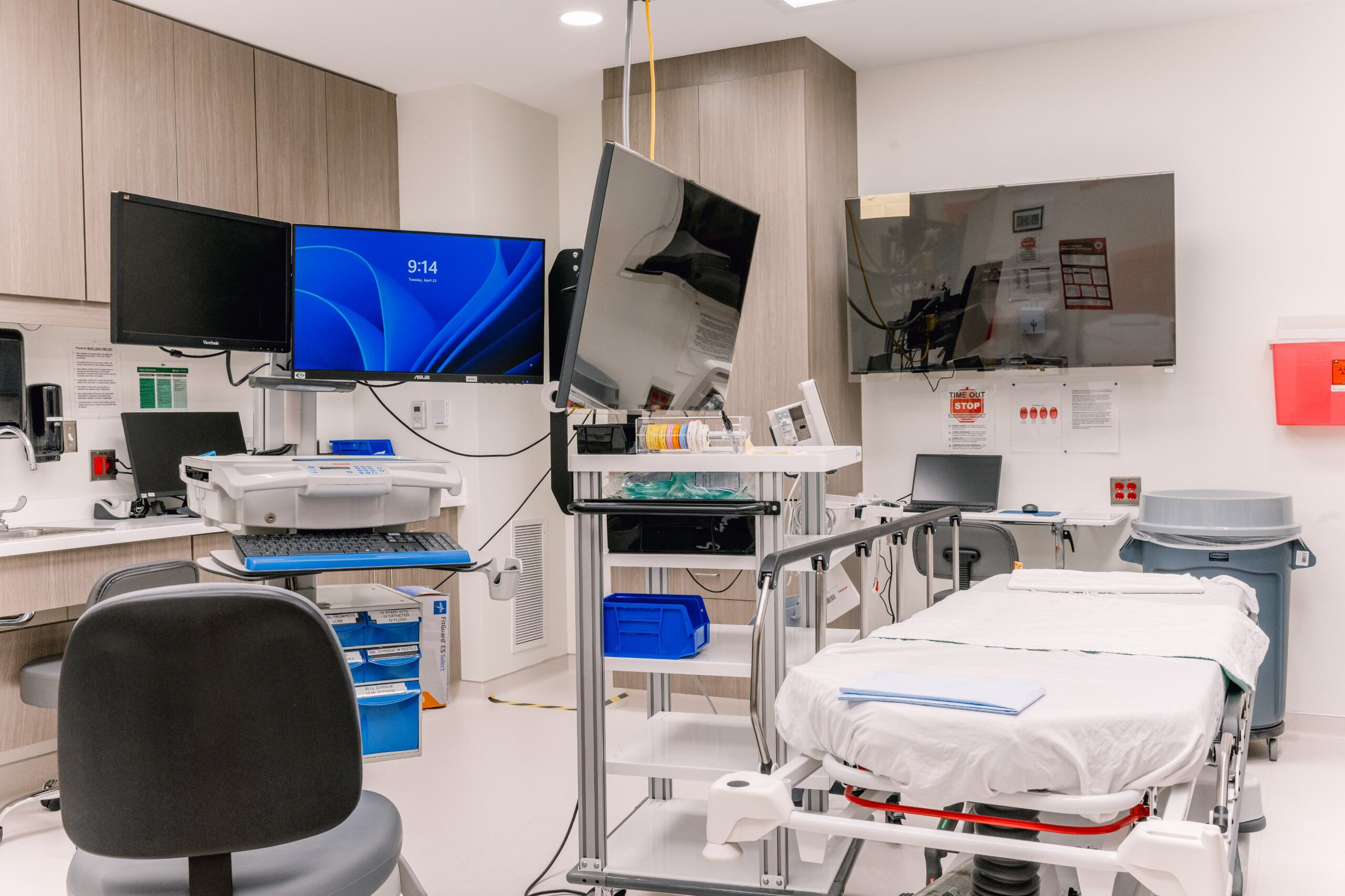Modern medical exam room with patient bed, equipment, monitors, printer, chairs, and cabinets; clean and organized space.