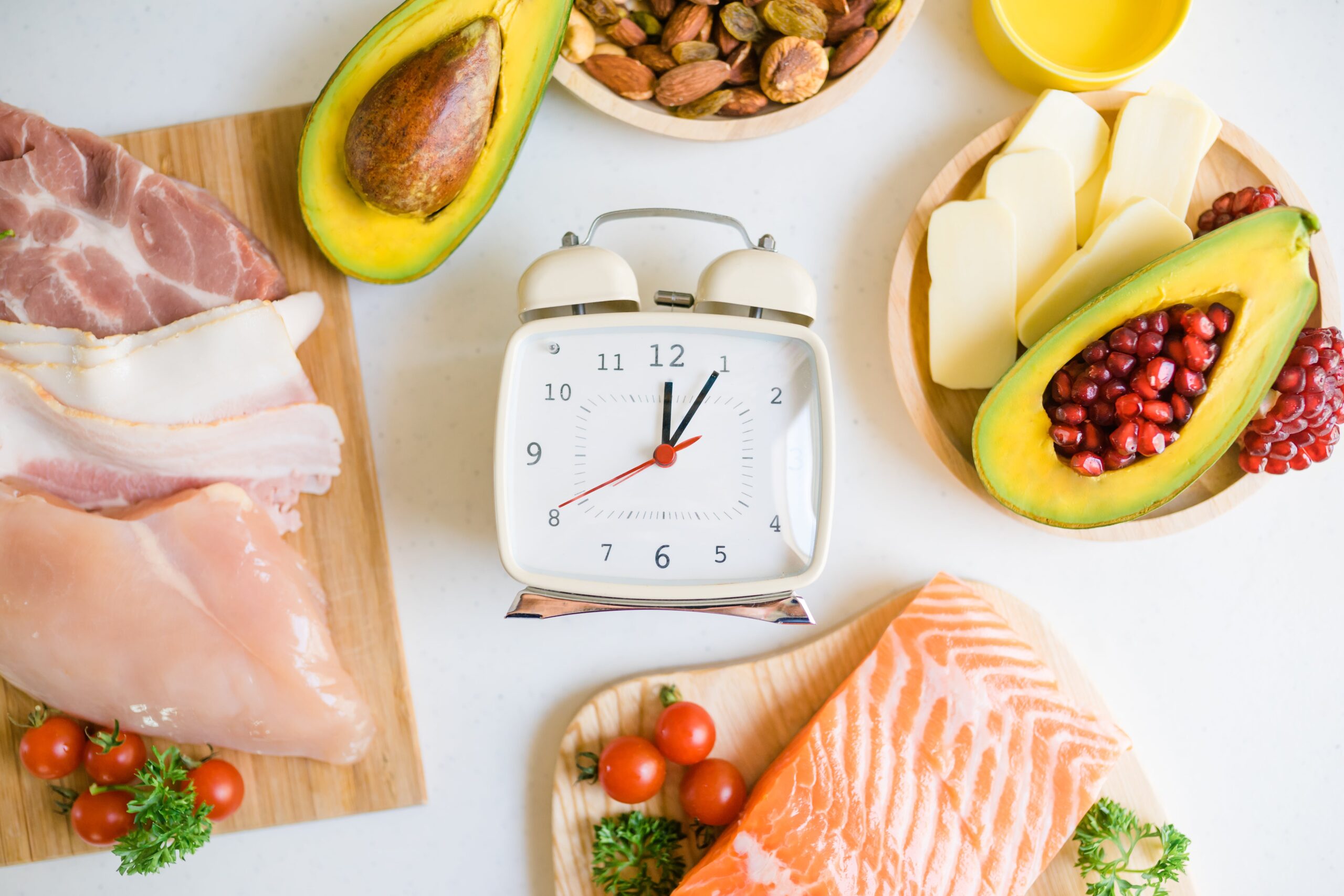 A white alarm clock with keto foods around it: salmon, chicken, bacon, ham, avocado halves, almonds, butter, tomatoes, pomegranate.