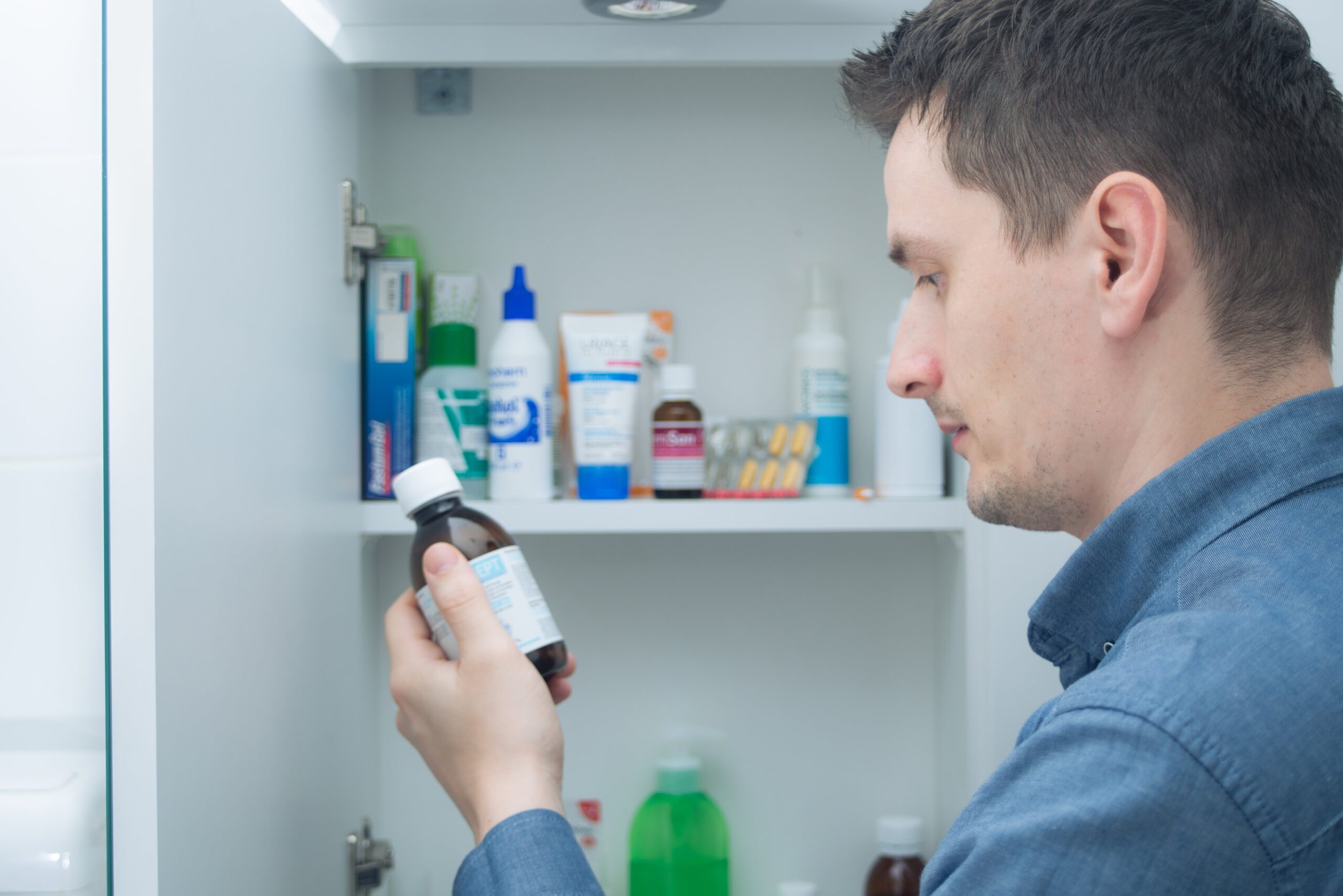 A man in a blue shirt looks at a medicine bottle in front of an open cabinet stocked with medications and health supplies.