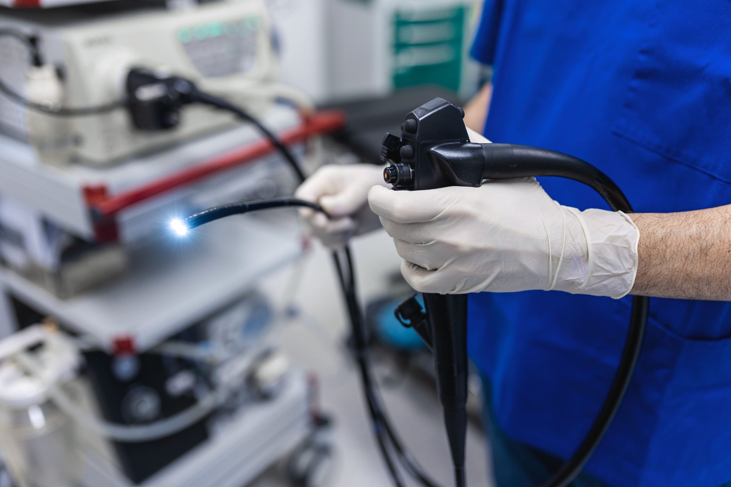 A medical professional in blue scrubs and gloves holds an illuminated endoscope beside medical equipment in a clinical environment.