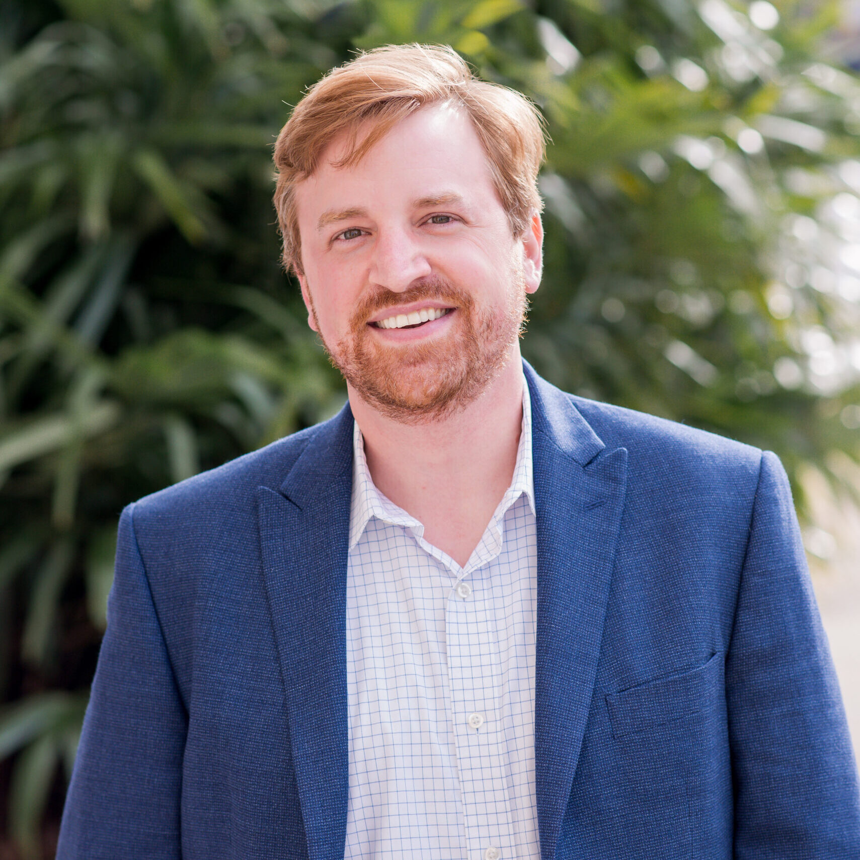 A man with light brown hair and a beard, in a blue blazer and checked shirt, smiles outdoors in front of green foliage.