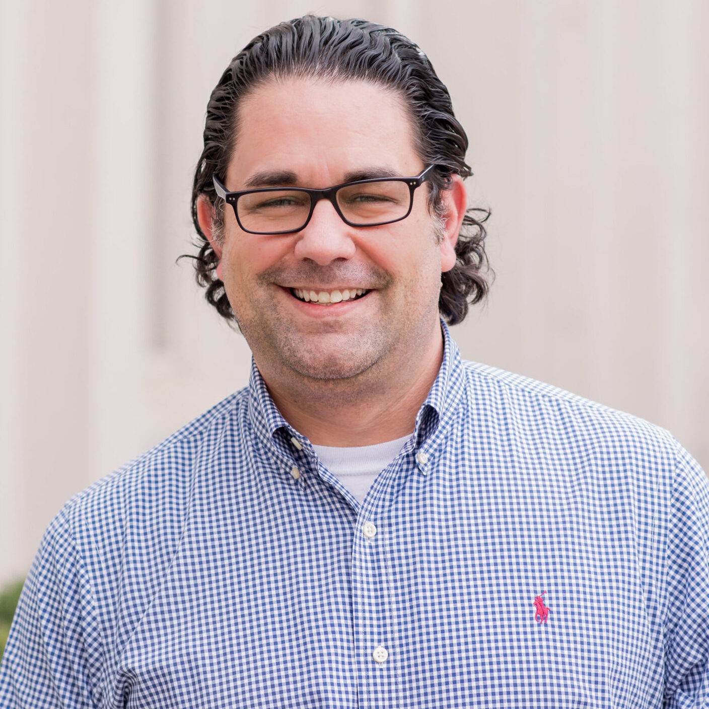 A man with dark wavy hair and glasses smiles at the camera, wearing a blue and white checkered shirt against a blurred background.