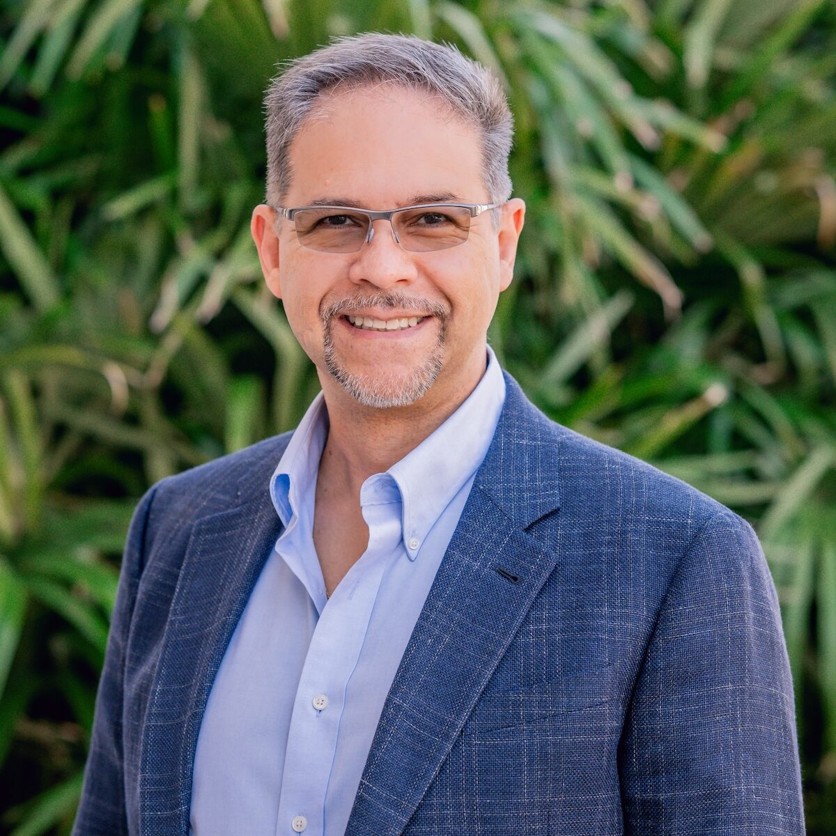 A man with glasses, dressed in a blue blazer and light blue shirt, smiles outdoors in front of vibrant green foliage.